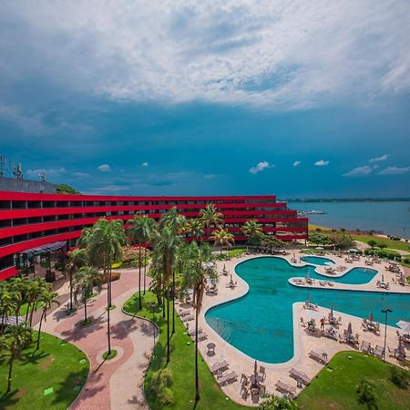 Royal Tulip Brasilia Alvorada Hotel Kültér fotó The photo shows a modern hotel building with a striking red exterior, surrounded by lush greenery. In the foreground, there is a large, creatively shaped swimming pool, with lounge chairs arranged around it. The backdrop features a body of water, pos