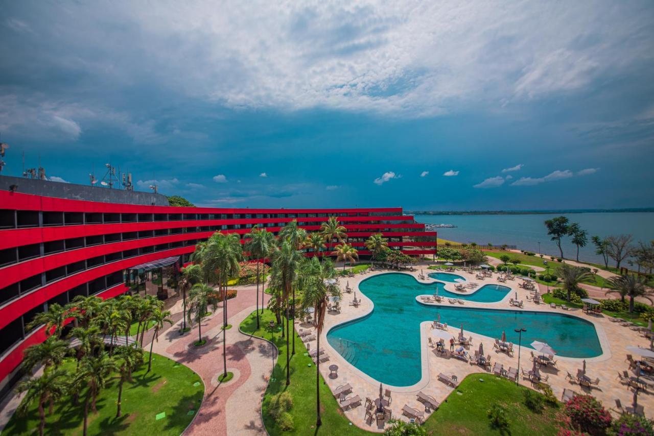 Royal Tulip Brasilia Alvorada Hotel Kültér fotó The photo shows a modern hotel building with a striking red exterior, surrounded by lush greenery. In the foreground, there is a large, creatively shaped swimming pool, with lounge chairs arranged around it. The backdrop features a body of water, pos