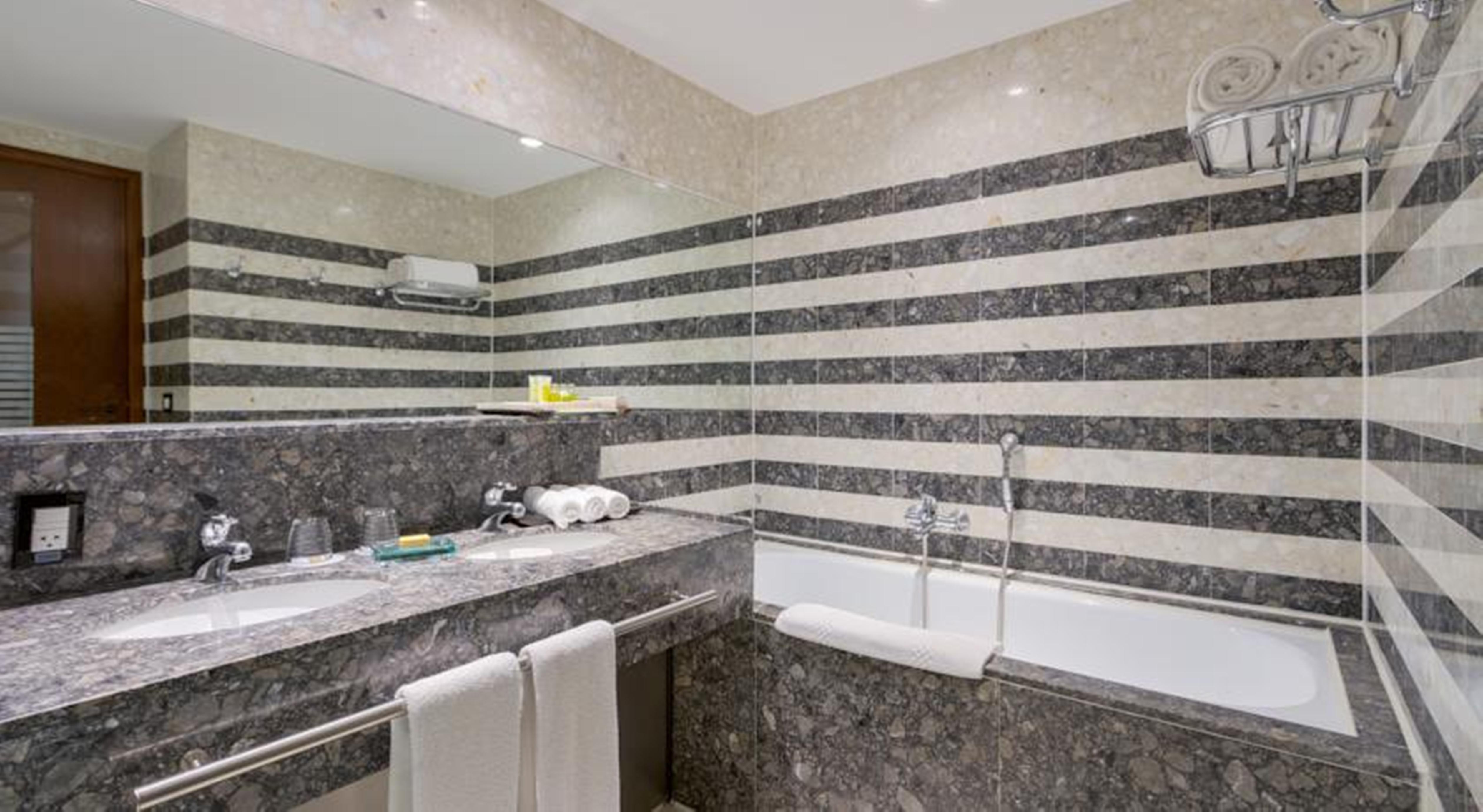 Royal Tulip Brasilia Alvorada Hotel Kültér fotó The photo shows a modern bathroom featuring a combination of marble and stone finishes. The walls are decorated with horizontal stripes in various shades of gray and cream. There is a bathtub with a white exterior, and a showerhead is mounted on the 