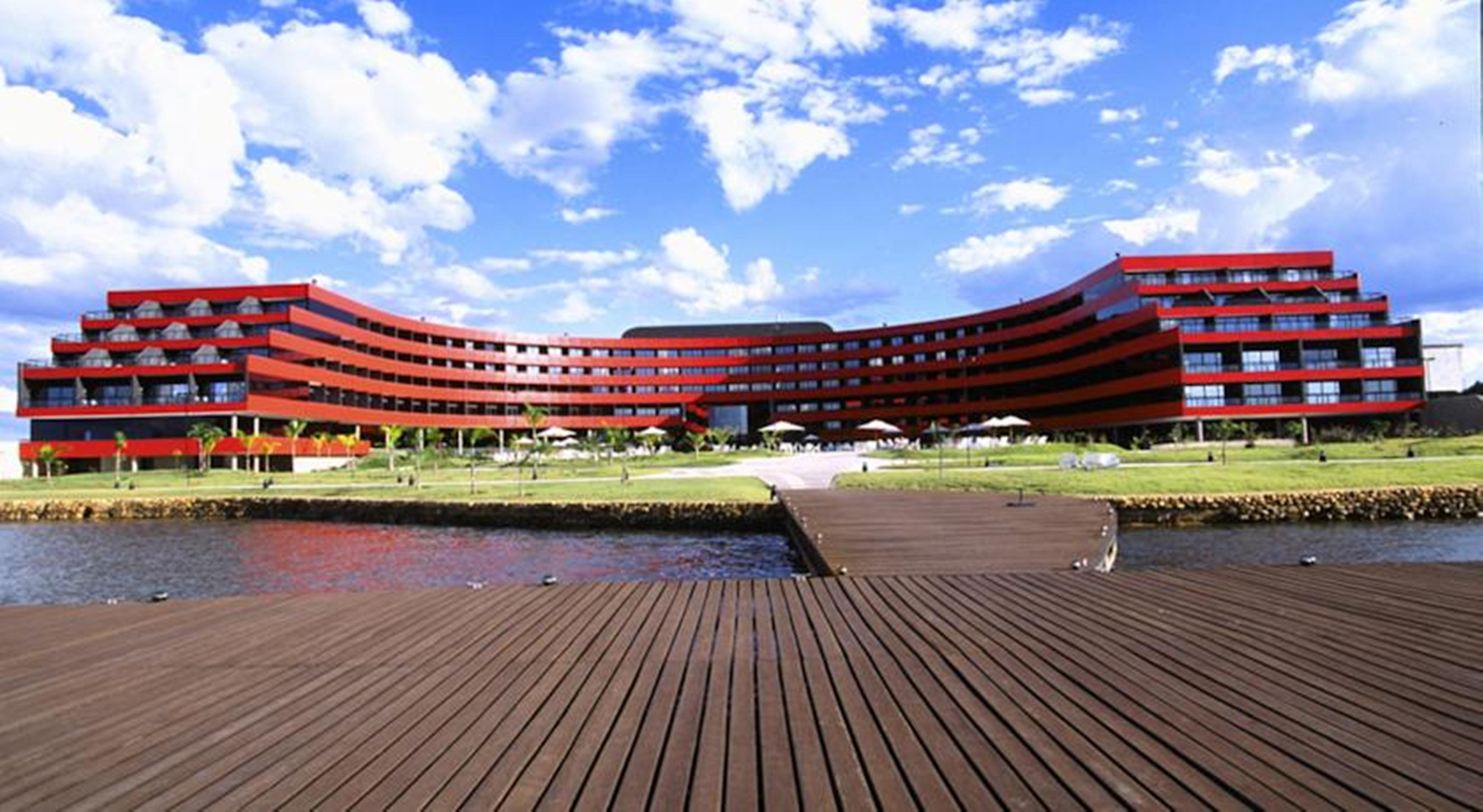Royal Tulip Brasilia Alvorada Hotel Kültér fotó The image shows a modern, curved building with a striking red facade, situated near a body of water. The architecture features multiple levels with large windows that create a sleek and contemporary appearance. In front of the building, there is a wo