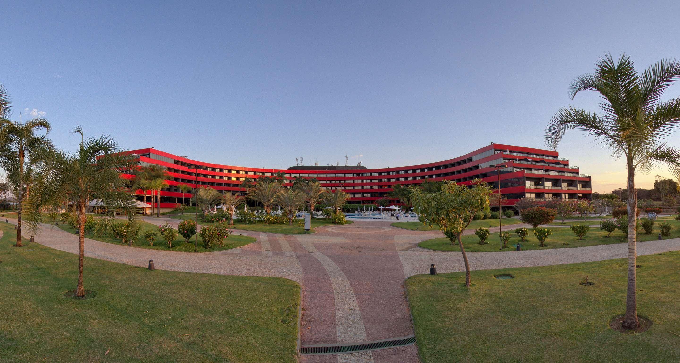 Royal Tulip Brasilia Alvorada Hotel Kültér fotó The photo shows a large, curved building with a vibrant red exterior. It appears to be a hotel or resort, surrounded by well-maintained gardens and green spaces. The landscape includes manicured lawns, small trees, and pathways, suggesting a pleasant