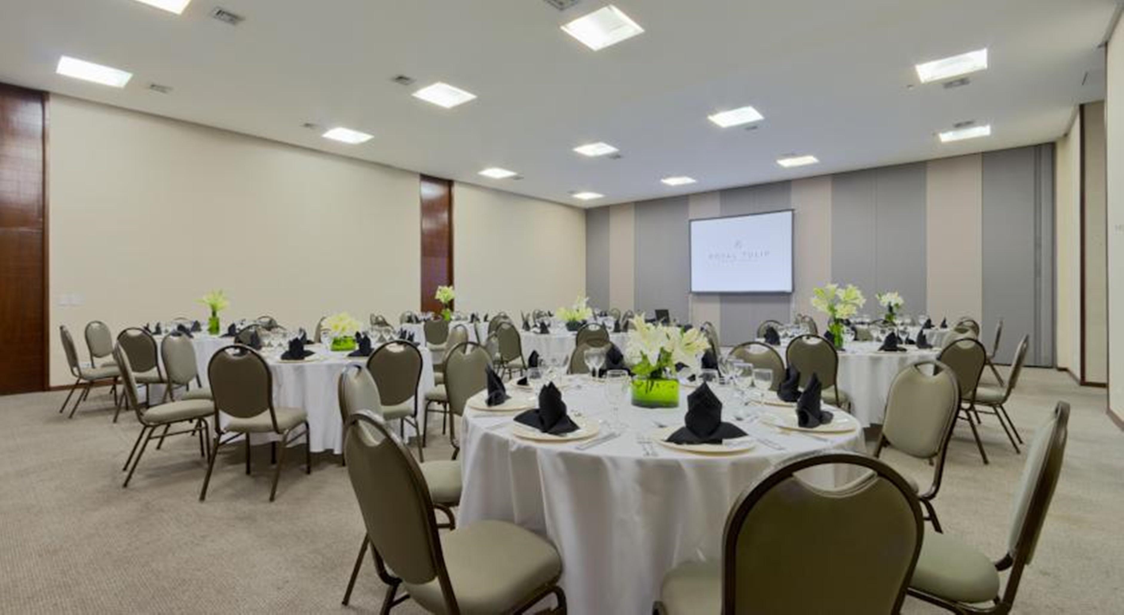 Royal Tulip Brasilia Alvorada Hotel Kültér fotó The photo shows a banquet or conference room set up with multiple round tables covered in white tablecloths. Each table is arranged with black napkins and centerpieces featuring flowers, likely in shades of green and white. The room has beige walls a
