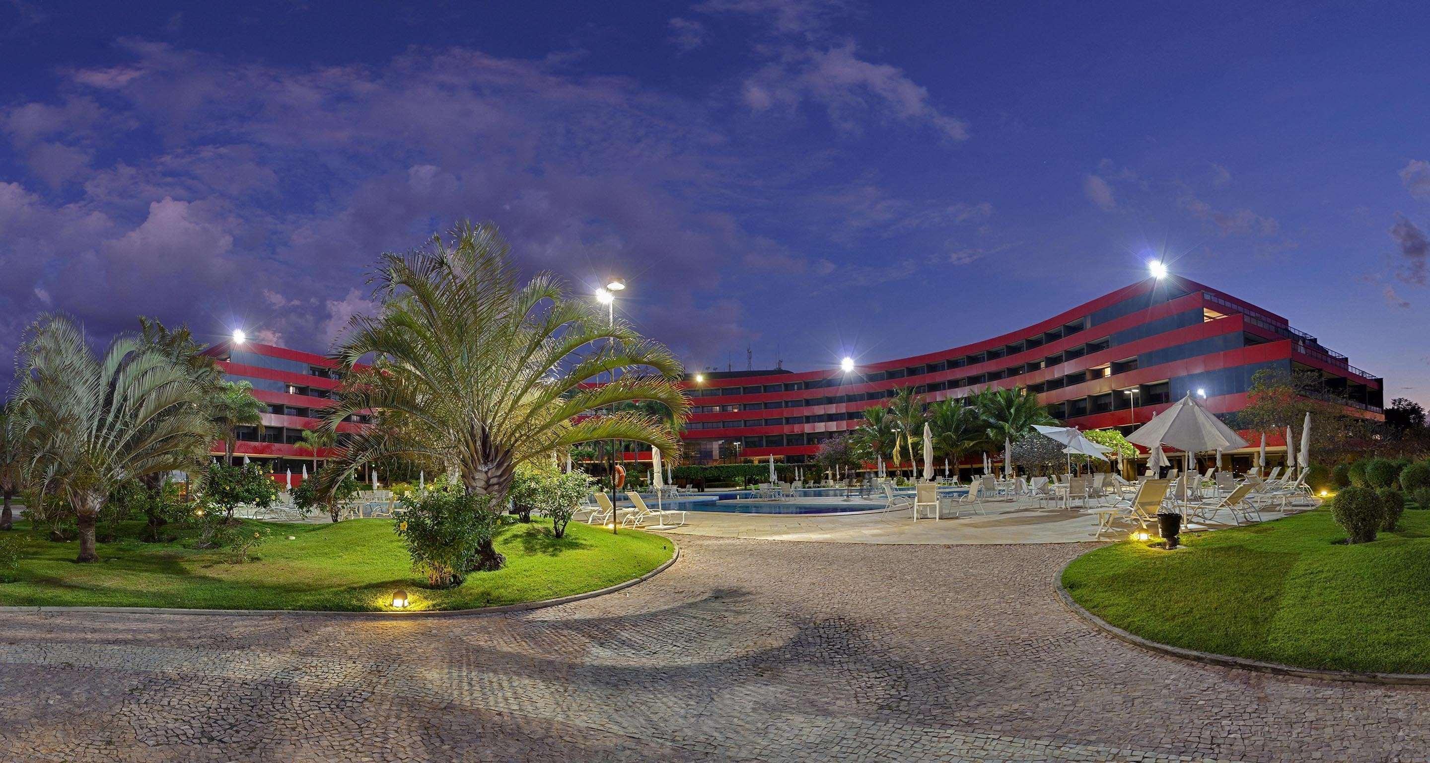 Royal Tulip Brasilia Alvorada Hotel Kültér fotó The photo shows a panoramic view of a hotel or resort complex, characterized by its modern architectural design with smooth, curved lines and a vibrant red color. It’s set in a landscaped area featuring palm trees and green grass. The surroundings in