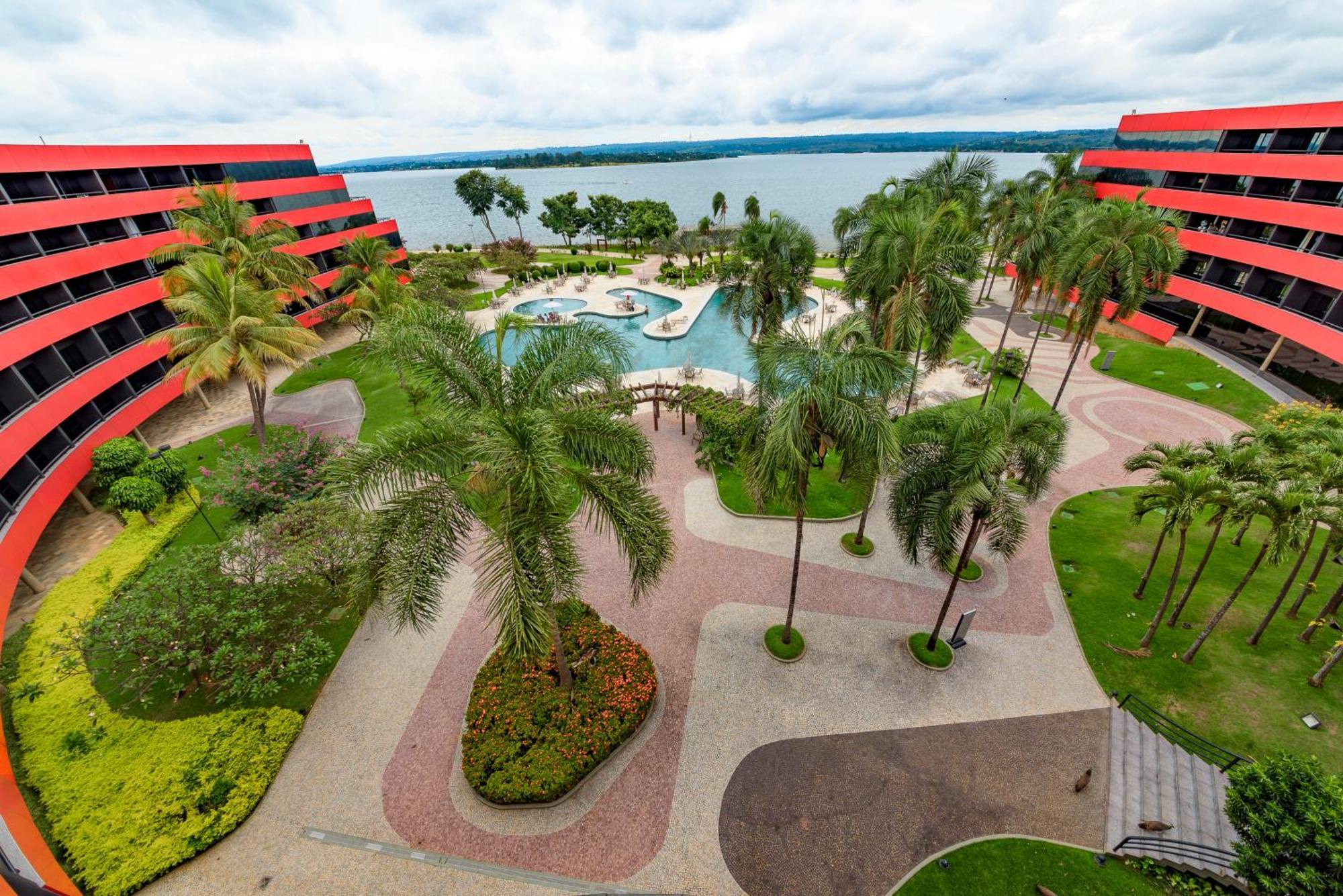 Royal Tulip Brasilia Alvorada Hotel Kültér fotó The photo shows a scenic view of a courtyard area in a residential complex. There are multiple red buildings surrounding a pool area, which is surrounded by palm trees and landscaped gardens. The pool features a curvy design, and there are pathways m