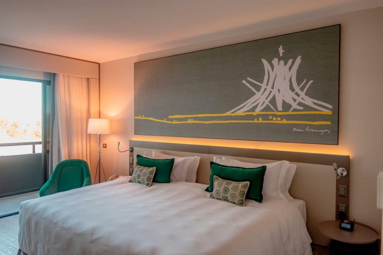 Royal Tulip Brasilia Alvorada Hotel Kültér fotó The photo shows a modern hotel room featuring a large bed with white bedding and two green accent pillows. Above the bed, there is a large piece of artwork depicting a stylized mountain or volcanic shape with yellow accents. The room has a contempora